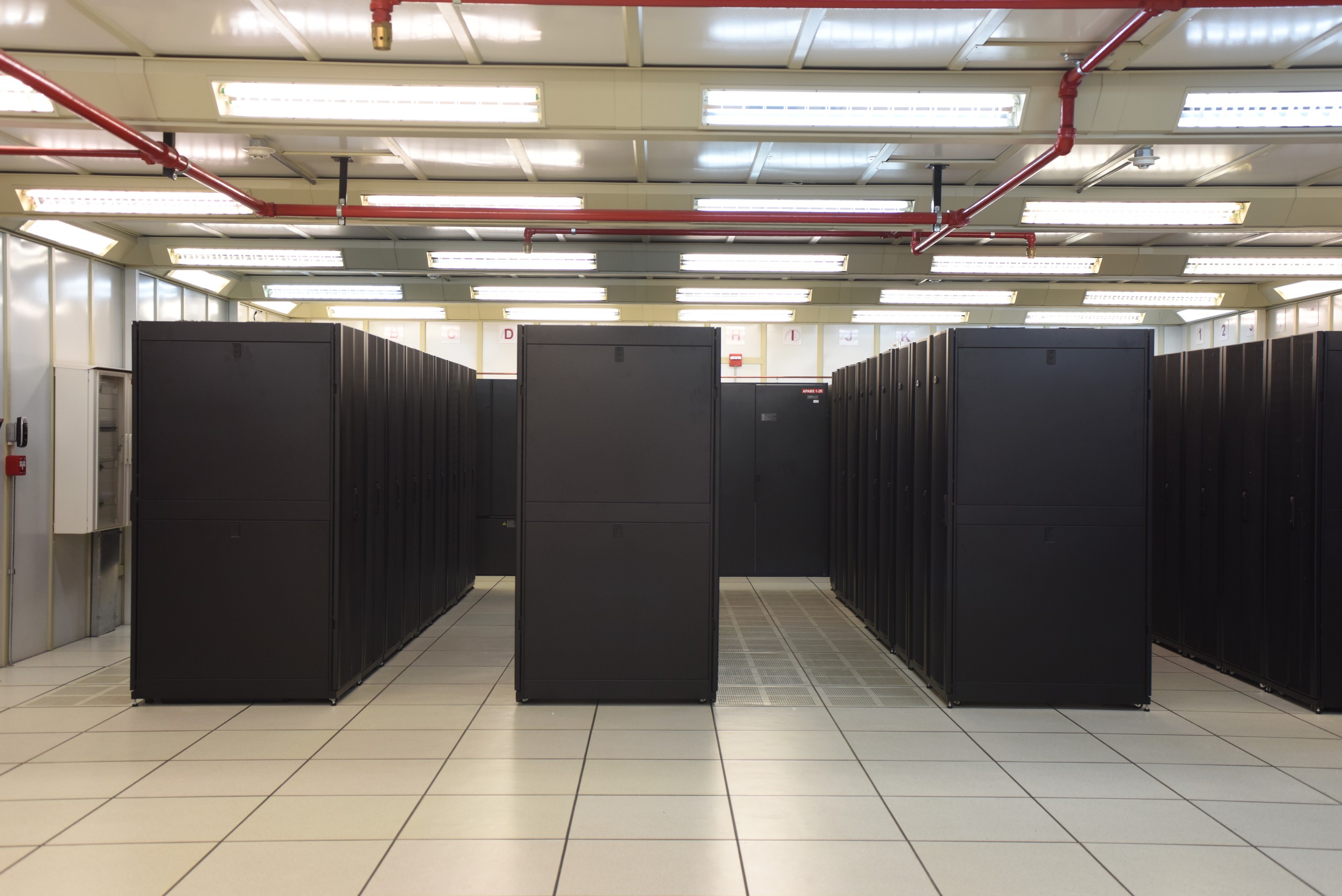 Rows of black server cabinets on a raised floor in a large data hall with overhead cable trays